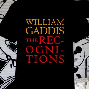 May include: Black t-shirt with the text "WILLIAM GADDIS THE RECOGNITIONS" in gold and red lettering. The shirt is displayed against a backdrop of a black and white mural.