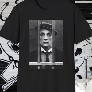May include: Black t-shirt with a black and white image of a person behind bars, wearing a hat and tie. The image is a vintage portrait. The shirt is made of a soft material.