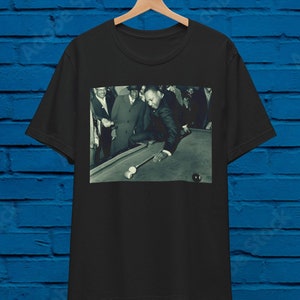 May include: A black and white photo of a man playing pool. He is wearing a suit and tie and is leaning over the pool table. There are other people in the background.