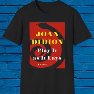 May include: Black t-shirt with a red rectangle design. The text reads "JOAN DIDION" in yellow, "Play It as It Lays" in white, and a black snake illustration. The words "A Novel" are at the bottom.