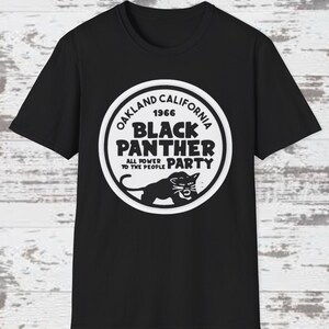 Può includere: T-shirt nero con grafica circolare bianca. La grafica include il testo "OAKLAND CALIFORNIA 1966 BLACK PANTHER PARTY ALL POWER TO THE PEOPLE" e un'illustrazione di pantera. La maglietta è realizzata in materiale morbido.