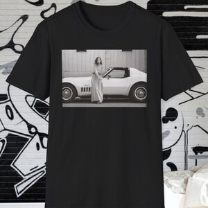May include: Black t-shirt featuring a black and white photograph. The photograph shows a person in a long dress standing next to a white sports car. The car is parked in front of a building.