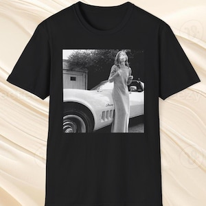 May include: Black t-shirt with a monochrome image of a woman in a long dress beside a white vintage car. The photograph has a classic, vintage aesthetic.