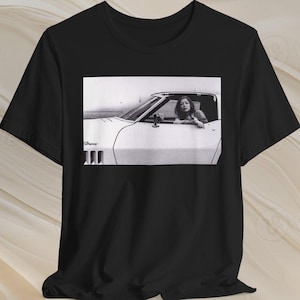 May include: Black t-shirt with a monochrome image of a woman in a white convertible. The car is a classic model, with the woman looking out the window. The image is printed on the front of the shirt.