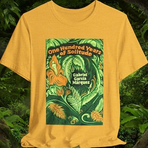 Puede incluir: Camiseta de color amarillo mostaza con el gráfico de la portada del libro "Cien años de soledad" de Gabriel García Márquez. El gráfico es una ilustración colorida con tonos naranja y verde. La camiseta es de tela suave.