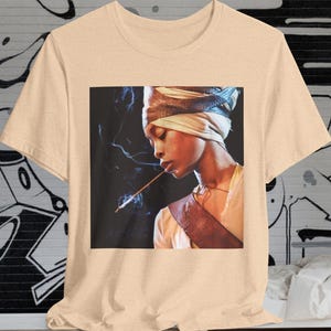 Op de afbeelding: Een beige T-shirt met een grafische print van een vrouw die een tulband draagt en een sigaret vasthoudt. De vrouw kijkt naar beneden en heeft een serieuze uitdrukking op haar gezicht.