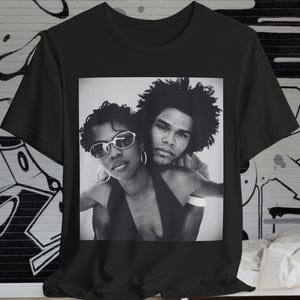 Può includere: T-shirt nero con una fotografia in bianco e nero di due persone che si abbracciano. La fotografia mostra una donna con occhiali da sole e orecchini a cerchio e un uomo con i capelli ricci. La t-shirt è esposta su un muro bianco con graffiti.