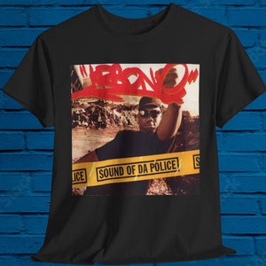 以下が含まれることがあります： 赤い文字で「FEENO」と書かれたスプレー缶を持った、野球帽とサングラスをかけた男性のグラフィックがプリントされた黒いTシャツ。グラフィックは、「SOUND OF DA POLICE」という文字が書かれた黄色と黒の警察用テープで囲まれています。