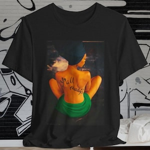 Puede incluir: Camiseta negra con un estampado gráfico de una mujer de espaldas. La mujer lleva una falda verde y tiene las palabras "y'all ready?" escritas en la espalda.