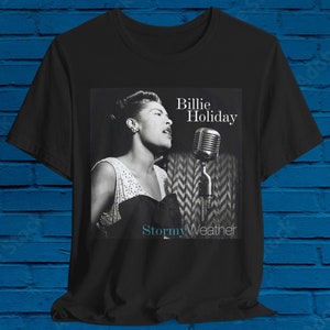 Può includere: Foto in bianco e nero di Billie Holiday che canta in un microfono. Il testo "Billie Holiday" e "Stormy Weather" sono stampati sulla t-shirt.