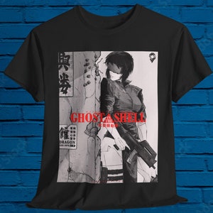 以下が含まれることがあります： 銃を持った女性のグラフィックプリントが施された黒いTシャツ。グラフィックは白黒で、赤い文字で「攻殻機動隊」と書かれています。