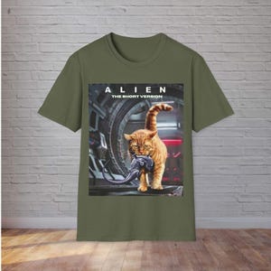 May include: Khaki green t-shirt with a graphic of a ginger cat carrying an alien. The text "ALIEN THE SHORT VERSION" is above the cat. The shirt is displayed against a brick wall.