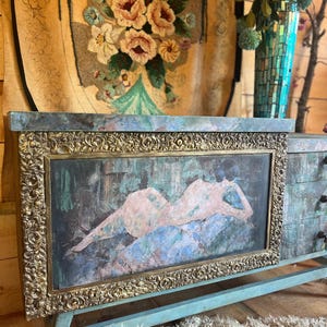 May include: A vintage turquoise blue dresser with a gold ornate frame around a painting of a nude figure. The dresser has three drawers and sits on a wooden floor.