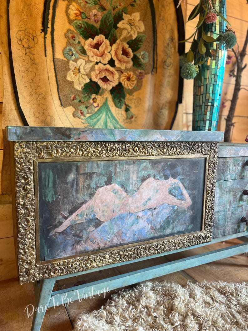 May include: A vintage turquoise blue dresser with a gold ornate frame around a painting of a nude figure. The dresser has three drawers and sits on a wooden floor.