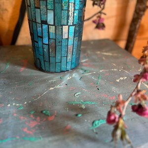 May include: A teal blue mosaic glass vase with a speckled gray surface. The vase is sitting on a gray surface with splashes of green, red, and white paint.