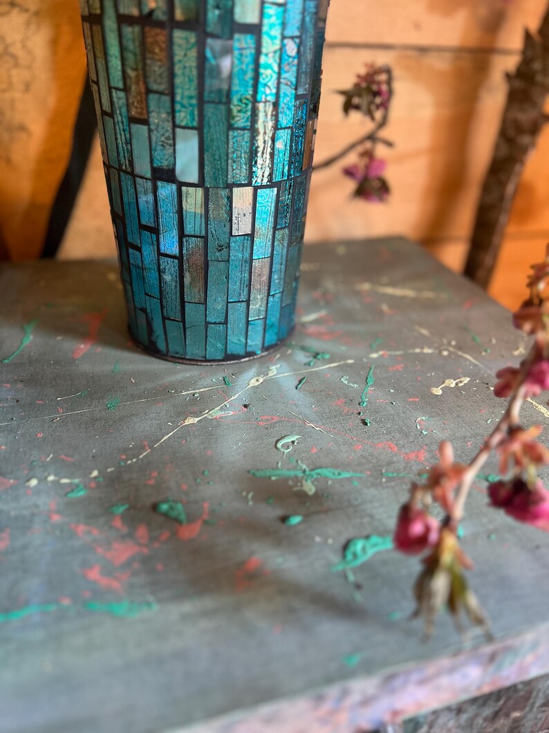 May include: A teal blue mosaic glass vase with a speckled gray surface. The vase is sitting on a gray surface with splashes of green, red, and white paint.