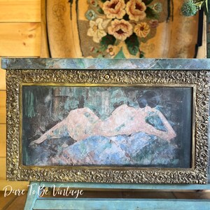 May include: A vintage wooden chest with a teal blue paint finish and gold accents. The chest features a decorative brass frame with an ornate design. The front panel of the chest has a painted image of a nude figure reclining on a patterned background. The chest is sitting on a rug with a wooden floor in the background.