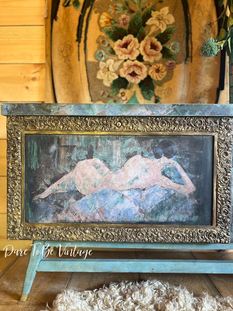 May include: A vintage wooden chest with a teal blue paint finish and gold accents. The chest features a decorative brass frame with an ornate design. The front panel of the chest has a painted image of a nude figure reclining on a patterned background. The chest is sitting on a rug with a wooden floor in the background.