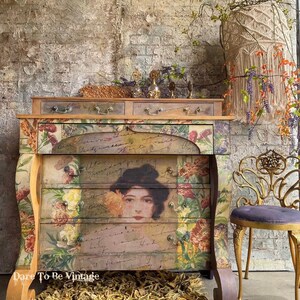 May include: A vintage-style dresser with floral and handwritten designs. The dresser has four drawers and a top shelf. The dresser is painted in a distressed white finish and has gold accents. The dresser is sitting on a rug made of natural fibers. The dresser is decorated with a floral arrangement and a small, gold-colored chair.