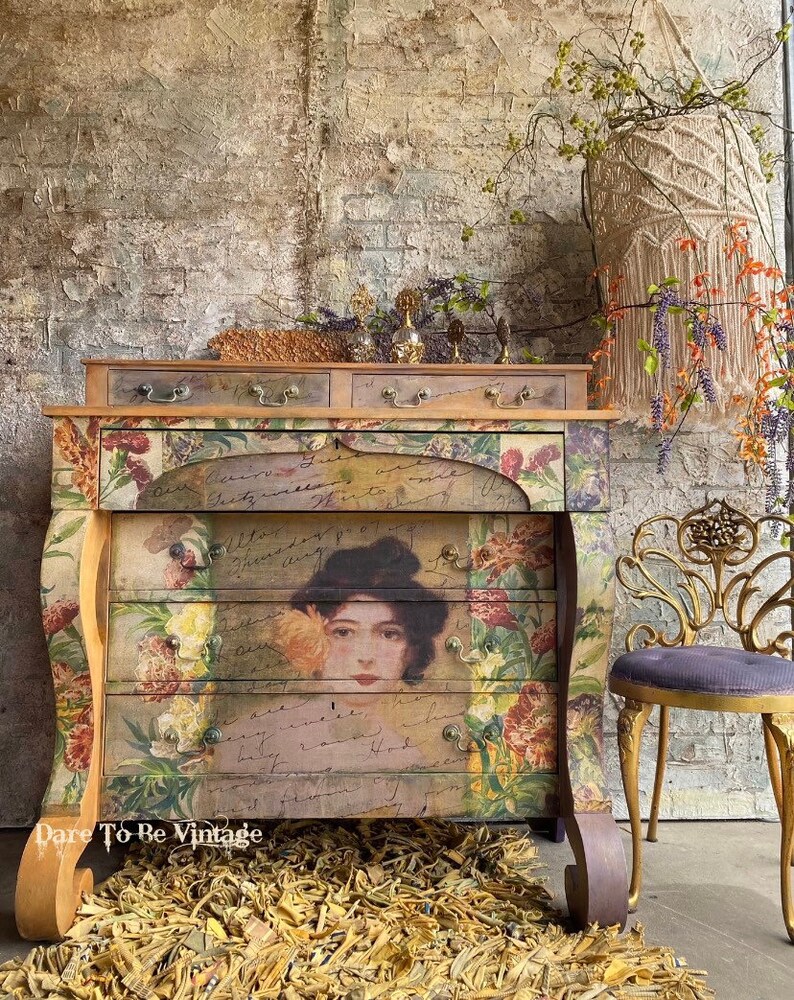 May include: A vintage-style dresser with floral and handwritten designs. The dresser has four drawers and a top shelf. The dresser is painted in a distressed white finish and has gold accents. The dresser is sitting on a rug made of natural fibers. The dresser is decorated with a floral arrangement and a small, gold-colored chair.