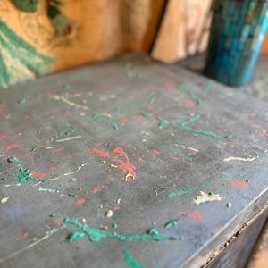 May include: A gray wooden box with a distressed finish. The box is decorated with splashes of green, pink, and white paint.