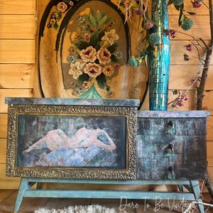 May include: A teal blue vintage dresser with three drawers and a cabinet door. The cabinet door has a gold frame and a framed abstract painting of a nude figure. The dresser is sitting on a white fluffy rug. The dresser is decorated with a turquoise mosaic vase with pink flowers.