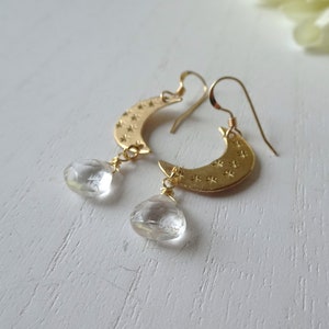 May include: Gold crescent moon earrings with clear quartz teardrop dangles. The crescent moon is textured with small stars.