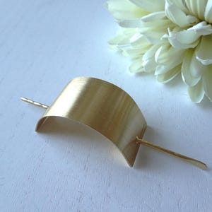 May include: A gold-colored hair barrette with a curved design and a straight pin. The barrette is made of metal and has a smooth, polished finish. A white flower is in the background.