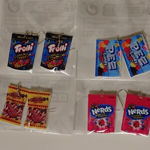 May include: A set of six pairs of earrings featuring popular candy brands. The earrings are made of polymer clay and feature images of Trolli Sour Brite Crawlers, Nerds Gummy Clusters, and Pez Candy.