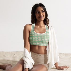 May include: A woman wearing a green sports bra with a white trim and a white and orange floral pattern. She is sitting on a sandy beach with a white shirt draped over her shoulders.