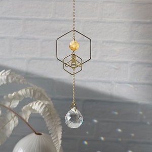 Hand holding a honeybee suncatcher with a bee pendant, hexagon, and citrine gemstone centerpiece, above a sparkling prism against a white brick backdrop.