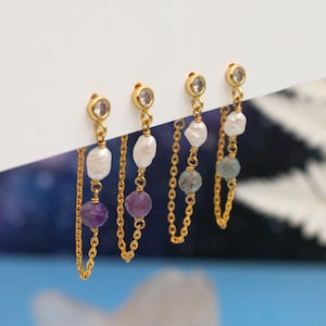 May include: Four pairs of gold-colored earrings with dangling chains and gemstones. Each earring features a small, clear stone at the top, followed by a chain with a gemstone and a pearl. The gemstones are purple, white, and blue-green.