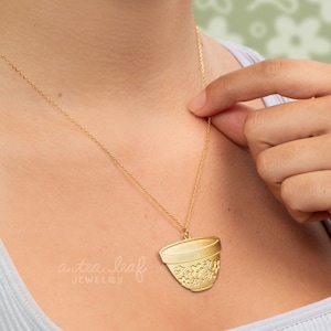 May include: A gold necklace with a pendant shaped like a teacup with a floral design. The pendant is hanging on a delicate gold chain. The text "a tea leaf jewelry" is visible in the image.