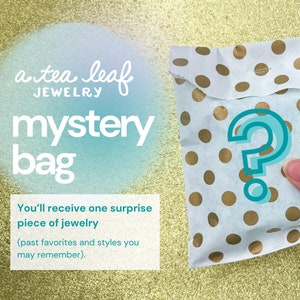 May include: A white paper bag with gold polka dots and a teal question mark. The bag is labeled "a tea leaf jewelry mystery bag". The text reads: "You'll receive one surprise piece of jewelry (past favorites and styles you may remember)."