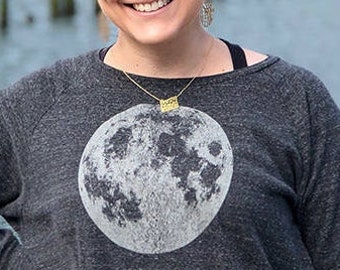 May include: A woman wearing a dark gray long-sleeved shirt with a white moon graphic. She is smiling and wearing a gold necklace with a small rectangular pendant.