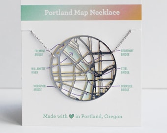 May include: A silver necklace with a map of Portland, Oregon, cut out of the metal. The map is surrounded by a circle and the necklace is on a white card with the text "Portland Map Necklace" and "Made with in Portland, Oregon".