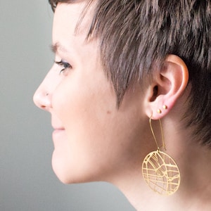 May include: A person with short brown hair wearing a gold geometric earring. The earring is a circle with a detailed design.