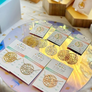 May include: A set of gold and silver earrings and necklaces featuring a map of Portland, Oregon. The jewelry is displayed on a white and pink surface with the text "a tea leaf jewelry" and "Portland Map Necklace".