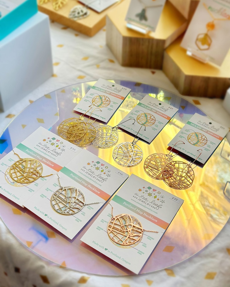 May include: A set of gold and silver earrings and necklaces featuring a map of Portland, Oregon. The jewelry is displayed on a white and pink surface with the text "a tea leaf jewelry" and "Portland Map Necklace".