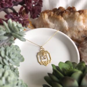 Puede incluir: Un collar de oro con un colgante geométrico que presenta un cristal transparente. El colgante tiene forma cuadrada con detalles intrincados. El collar se muestra en un plato blanco.