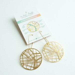 May include: Gold-toned earrings featuring a detailed map of Portland, Oregon. The earrings are circular and have a delicate wire design. The card reads "a tea leaf jewelry" and "Portland Map Earrings".