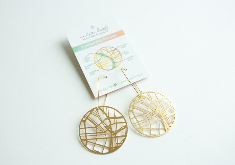 May include: Gold-toned earrings featuring a detailed map of Portland, Oregon. The earrings are circular and have a delicate wire design. The card reads "a tea leaf jewelry" and "Portland Map Earrings".