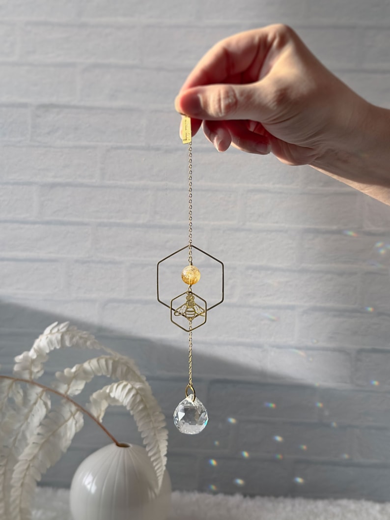 Hand holding a honeybee suncatcher with a bee pendant, hexagon, and citrine gemstone centerpiece, above a sparkling prism against a white brick backdrop.