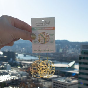 May include: Gold-toned earrings shaped like a map of Portland, Oregon. The earrings are hanging from a card that says "a tea leaf jewelry" and "Portland Map Earrings".