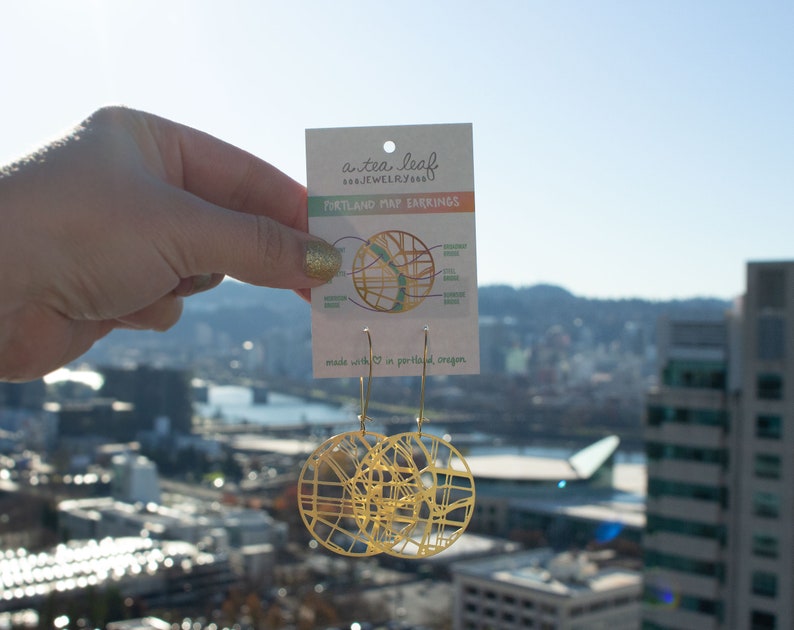 May include: Gold-toned earrings shaped like a map of Portland, Oregon. The earrings are hanging from a card that says "a tea leaf jewelry" and "Portland Map Earrings".