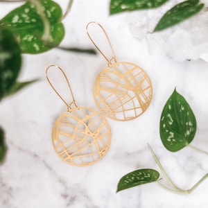 May include: A pair of gold-toned earrings with a circular design featuring a detailed map-like pattern. The earrings are hanging from a white surface.
