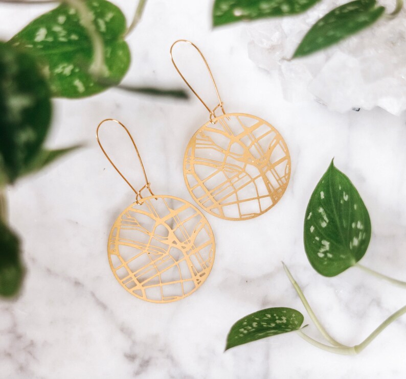 May include: A pair of gold-toned earrings with a circular design featuring a detailed map-like pattern. The earrings are hanging from a white surface.