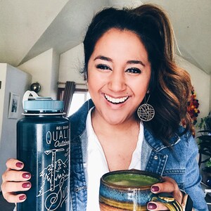 May include: A person holding a black water bottle with a white logo that says "REI CO-OP QUALITY Outdoors Since 1938" and a green matcha drink in a brown and blue ceramic mug.