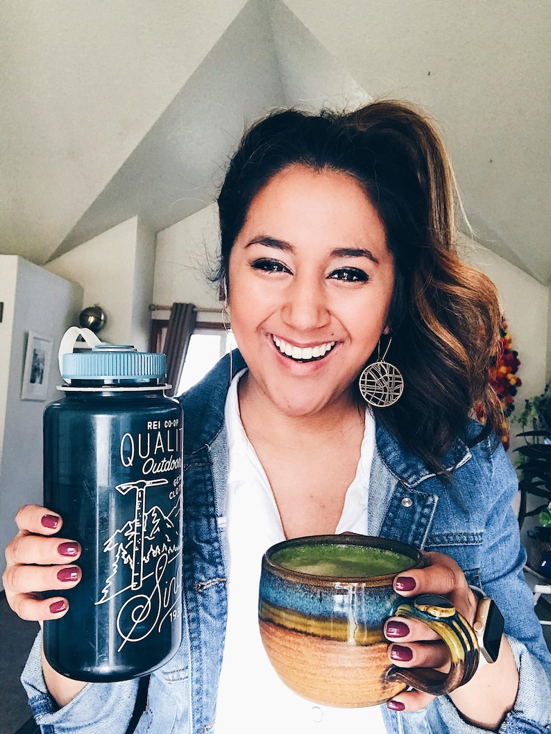 May include: A person holding a black water bottle with a white logo that says "REI CO-OP QUALITY Outdoors Since 1938" and a green matcha drink in a brown and blue ceramic mug.