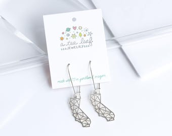 May include: A pair of silver-toned metal earrings shaped like the state of California with a geometric wireframe pattern. The earrings are attached to a white display card featuring the text a tea leaf JEWELRY and made with love in portland, oregon.